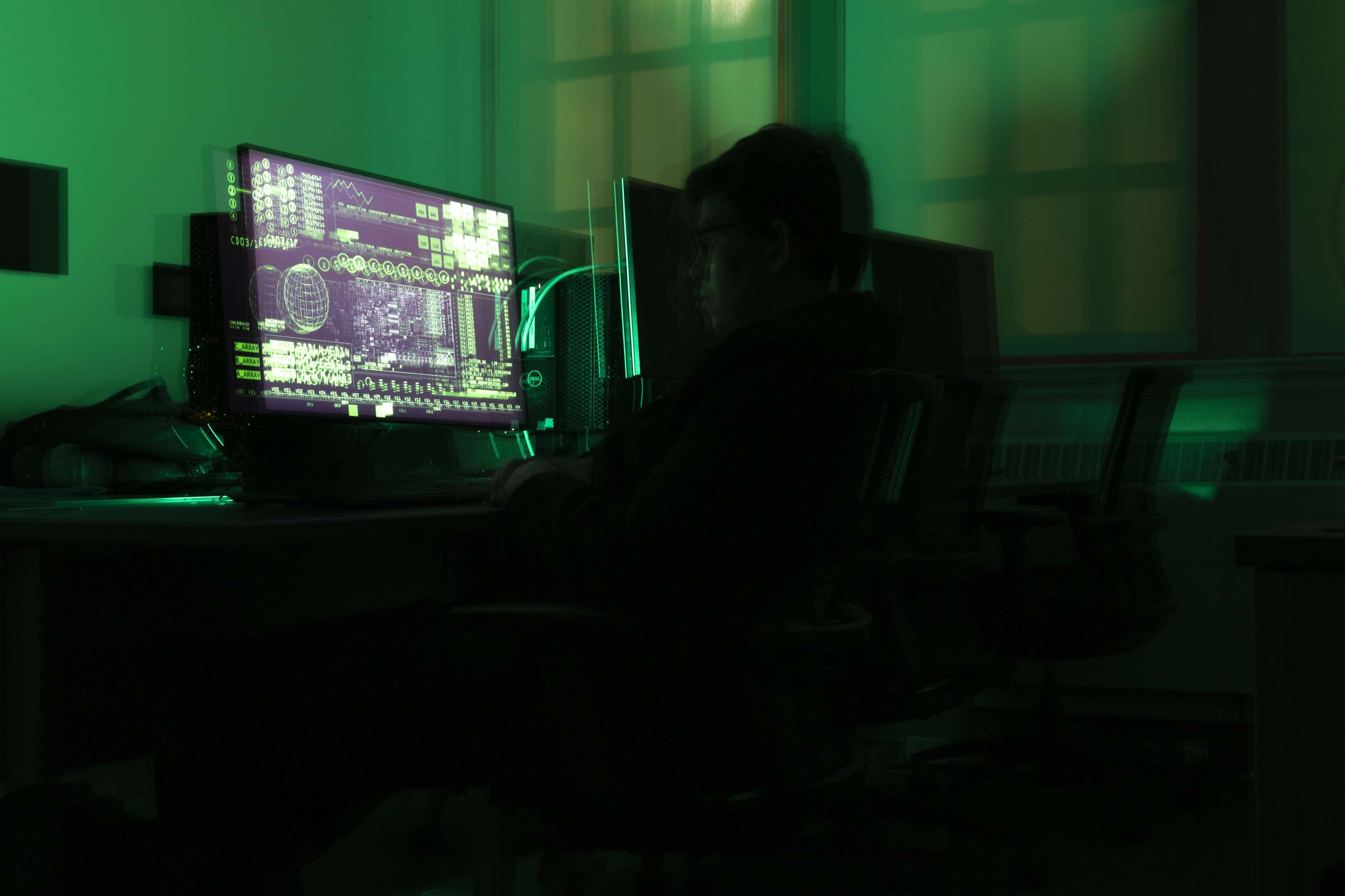 A person seated at a computer, with green light and projected code across the room.