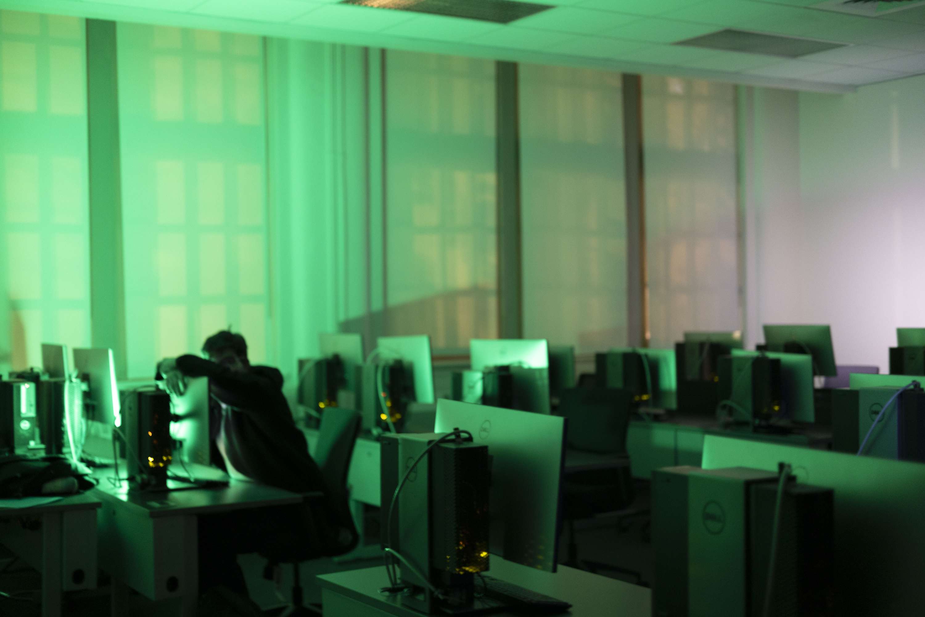 A person leaning forward at a desk among rows of computers in a green-lit room.