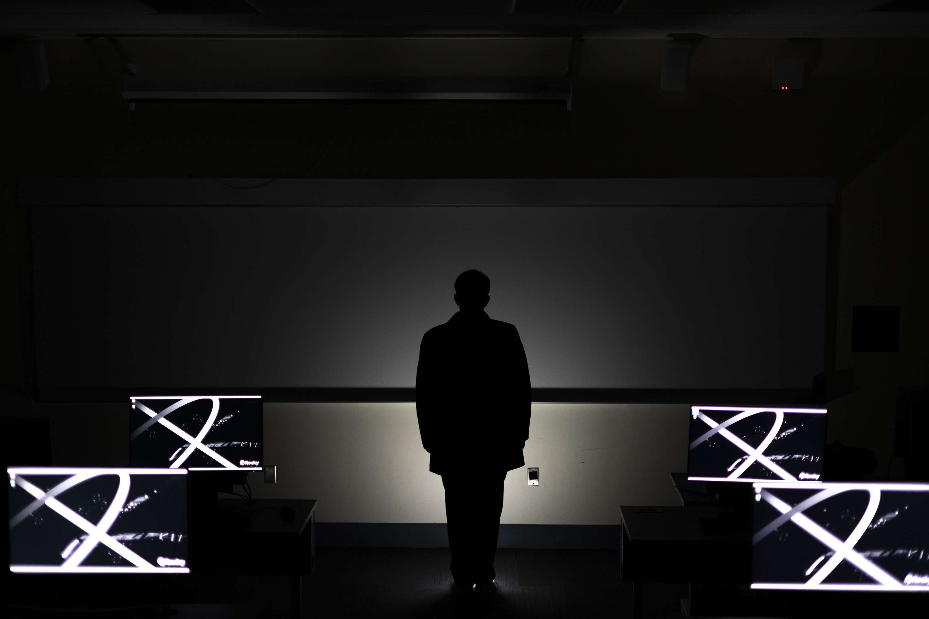 A standing silhouette between two rows of monitors and a softly illuminated wall.