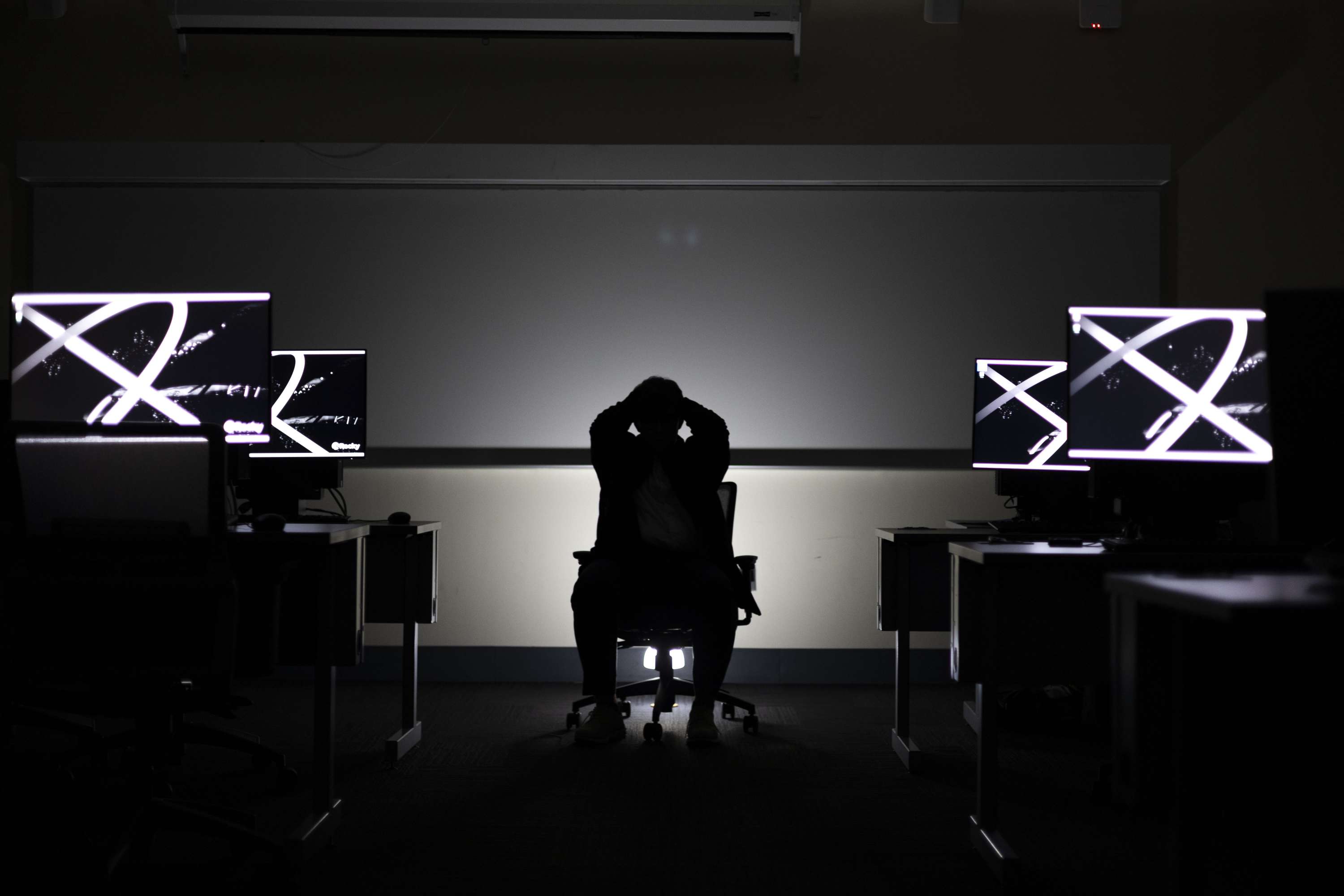A seated figure with hands raised to their head between symmetrical rows of glowing monitors.